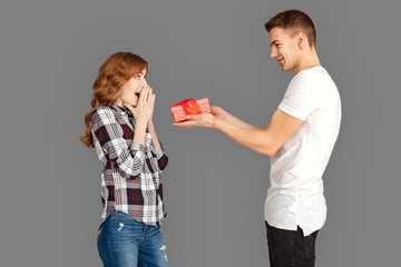 Celebration Concept. Young boyfriend giving present to surprised girlfriend standing isolated on grey