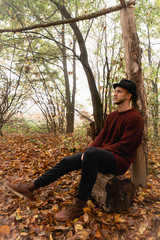 Young stylish man in black hat and red sweater sitting on the stump among the forest in fog and looking away