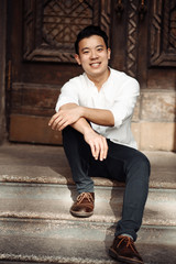 Portrait of handsome stylish asian guy in a white shirt and with a charming smile, who is sitting on the stairs, outdoors