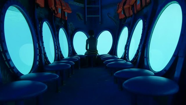 Push in on a boy sitting all alone facing away as he looks out the porthole of a submarine under a dreamlike blue glow.