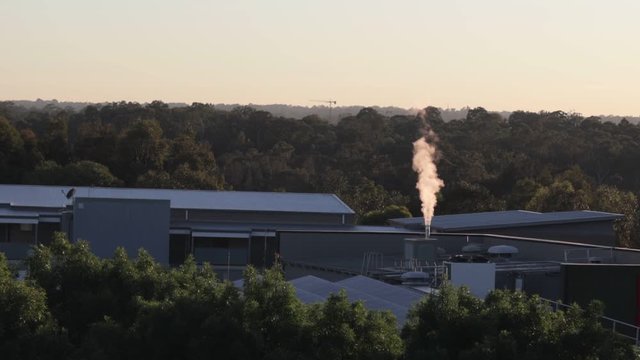 Steam and pollution coming out of industrial factory chimney creating environmental smoke and emissions in the tranquil Australian bush and residential development with a crane in the background.