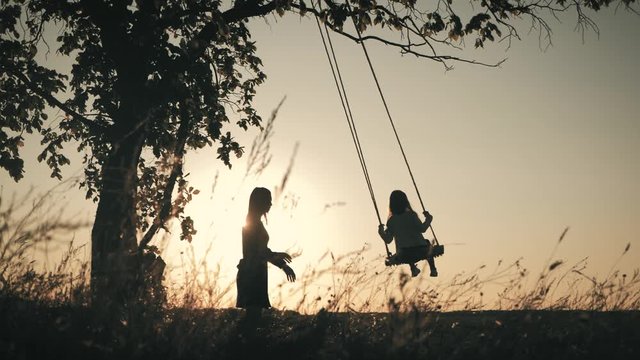 Silhouette of happy young mother and little daughter on a swing at sun light. Pretty girl sitting on a wooden swing and looking at golden sunset. Happy family concept.