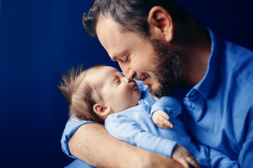 Middle age bearded Caucasian father hugging kissing newborn baby. Man parent holding child. Authentic lifestyle touching tender moment. Dad family life concept. Toned with classic blue 2020 colour. - obrazy, fototapety, plakaty