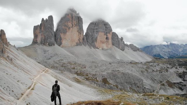 Aerial shot from a drone passing by a male model with backpack looking at Tre Cime di Lavaredo in the Dolomites, Italy