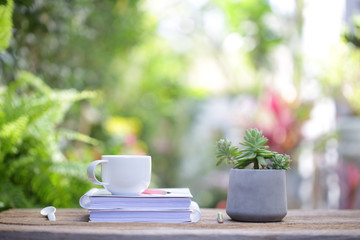 White coffee cup with small Succulent plant in old grey concrete pot with white thick book on wooden table at outdoor with nature bokeh background