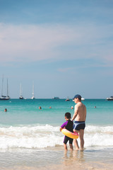 Dad and son in swimming suit holding hand then standing on the beach head to the sea with many yacht boats parking around in summer sunny day