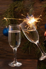 Glasses of champagne with bubbles on the background of Christmas decorations. Two glasses touch each other during a festive toast and sparklers burn beautifully. Beautiful card.