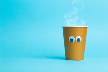 A paper cup with hot coffee and eyes on a blue background. Funny food concept