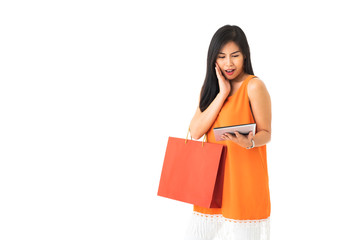 Fototapeta na wymiar A beautiful Asian young woman wearing orange dress holding a tablet and a shopping bag looking surprise in front of white isolated background.