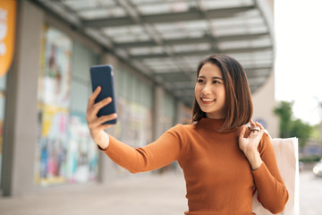 Beautiful young Asian woman holding phone to selfie with shopping bag outdoor shopping mall