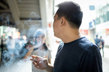 Asian man hold a cigarette,smokers father smoking near daughter,bad smell,pollution from cigarette smoke,smoking in a public place where people pass by in the city,dangerous to health,toxic to society © Satjawat