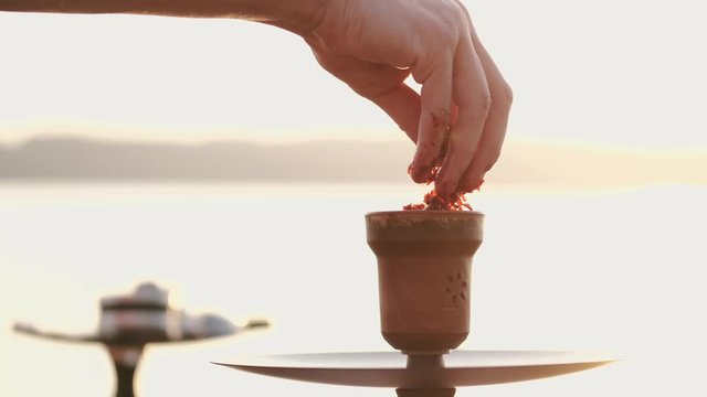 The hookah man is putting tobacco on the bowl. The sunset is on the background. The master is preparing hookah for vaping.