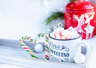 a white cup with marshmallows and Christmas sweets, on a white background with fir branches, balls and Christmas lights, as well as with a large red jar with deers in the background