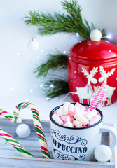 a white cup with marshmallows and Christmas sweets, on a white background with fir branches, balls and Christmas lights, as well as with a large red jar with deers in the background