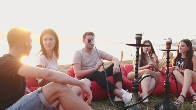 The circle of friends are smoking hookas and talking.The young are sitting in the beanbag chairs at sunset. They are sitting at the lakeside.