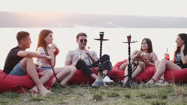 The circle of friends are smoking hookahs .The young are sitting in the beanbag chairs at sunset. They are sitting at the lakeside.
