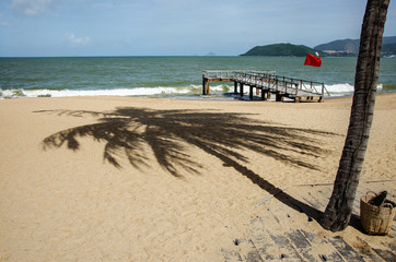 A shadow from a coconut tree falls on the sand. There is a pier for small boats on the beach. A red flag is posted on the coast - a ban on swimming. An island is visible on the horizon.