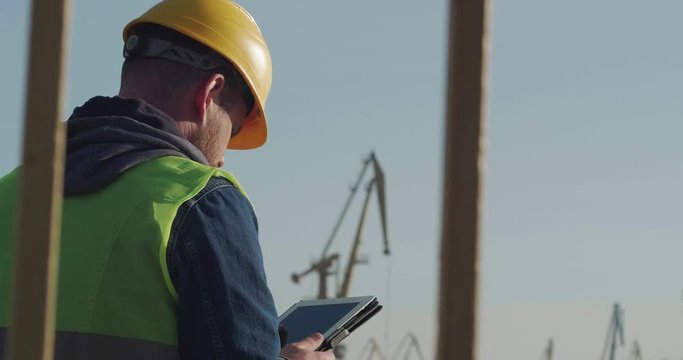A Dockworker With A Beard In A Yellow Helmet, Works With A Tablet PC In The Seaport Against The Background Of Cranes. The Foreman Inspects The Industrial Harbor. Cinema 4K Video