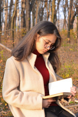 Obraz premium A girl in a light coat and glasses walks through the autumn Park with a book in her hands. The inscription on the book William Shakespeare. Soft focus.