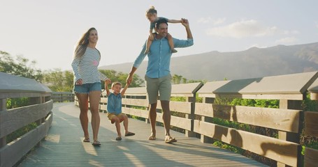 Happy smiling parents walking and swinging their young toddler son into the air, young toddler son riding on dads shoulders, family fun on a coastal boardwalk at sunset, weekend family walk - Powered by Adobe