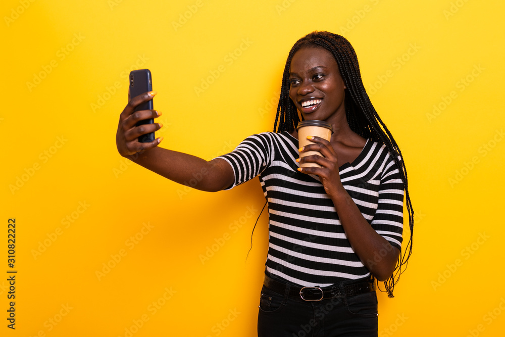 Wall mural Young african woman holding coffee cup take selfie on phone isolated on yellow background