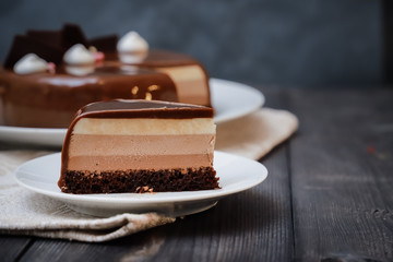 On a dark tabletop on a linen napkin, a tea composition: on a saucer a piece of cake made of three chocolate layers of different colors souffle, covered with glossy chocolate
