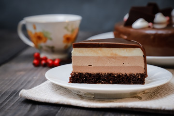 On a dark tabletop on a linen napkin, a tea composition: on a saucer a piece of cake made of three chocolate layers of different colors souffle, covered with glossy chocolate