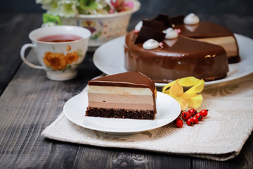 On a dark tabletop on a linen napkin, a tea composition: on a saucer a piece of cake made of three chocolate layers of different colors souffle, covered with glossy chocolate