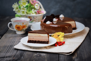 On a dark tabletop on a linen napkin, a tea composition: on a saucer a piece of cake made of three chocolate layers of different colors souffle, covered with glossy chocolate