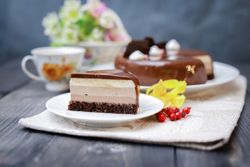 On a dark tabletop on a linen napkin, a tea composition: on a saucer a piece of cake made of three chocolate layers of different colors souffle, covered with glossy chocolate
