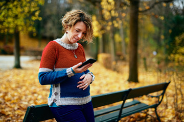 Pregnant blond woman in a park looking at her cell phone and smiling while holding her belly. The park is autumn yellow.