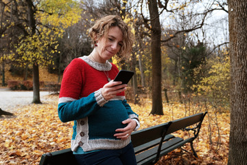 Pregnant blond woman in a park looking at her cell phone and smiling while holding her belly. The park is autumn yellow.