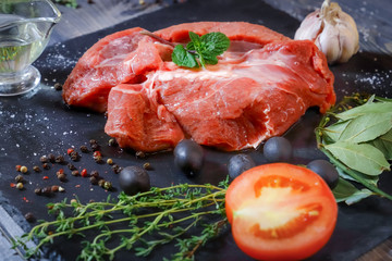 On a dark tabletop lies a piece of raw beef and spices bay leaf, thyme, mint, butter in a sauceboat