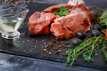 On a dark tabletop lies a piece of raw beef and spices bay leaf, thyme, mint, butter in a sauceboat
