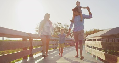 Happy smiling parents walking and swinging their young toddler son into the air, young toddler son riding on dads shoulders, family fun on a coastal boardwalk at sunset, weekend family walk - Powered by Adobe