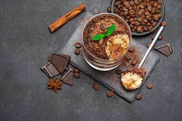 Classic tiramisu dessert in a glass cup and pieces of chocolate on stone cutting board on dark concrete background
