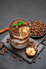 Classic tiramisu dessert in a glass cup and pieces of chocolate on stone cutting board on dark concrete background