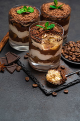 Classic tiramisu dessert in a glass cup and pieces of chocolate on stone cutting board on dark concrete background