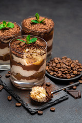 Classic tiramisu dessert in a glass cup and pieces of chocolate on stone cutting board on dark concrete background