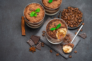 Classic tiramisu dessert in a glass cup and pieces of chocolate on stone cutting board on dark concrete background