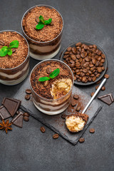 Classic tiramisu dessert in a glass cup and pieces of chocolate on stone cutting board on dark concrete background