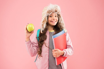 sweet and useful. Modern education. schoolgirl with books after lesson. school break. small happy girl earflap hat eat apple. winter holiday and vacation. teen kid pink background. pupil daily life