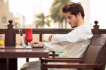 Young bearded businessman wearing Kandoura, sitting at table in cafe and uses laptop.Nearby is smartphone and cup of juice. Man checks time from his watch 