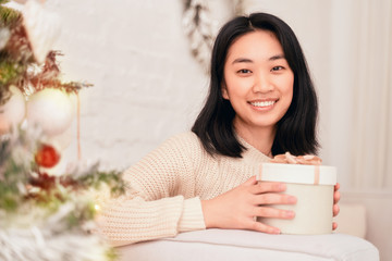 Girl fun looks at camera, holding gift for friend. Young woman of Asian appearance leaned on back of sofa against background of Christmas tree in home comfort of apartment, dressed in beige sweater.