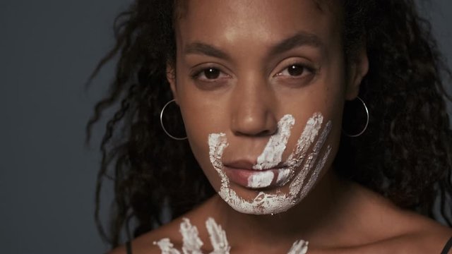 Serious calm african young woman with a handprint on her face and clavicles is standing with outstretched arm to the camera isolated over gray wall background
