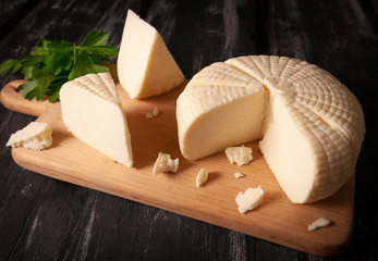 Suluguni cheese is cut into large pieces. On a light wooden board. Decorated with greenery. Dark...
