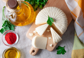 Suluguni cheese is cut into large pieces on a wooden board. Garnished with parsley leaves. Next to the cheese is a bottle with olive oil, parsley and a cup of coconut. On a gray linen background. 
