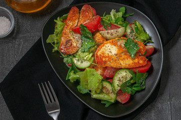 Salad with vegetables and fried halumi cheese with spices. On a black plate. Next to the plate is a fork and jars of spices with butter. View from above. Dark background backdrop.