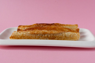 Fried pancakes roll into a tube on a white plate with a pink background.