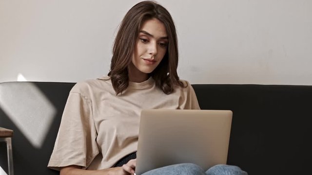 Smiling brunette woman using laptop computer while sitting on sofa indoors